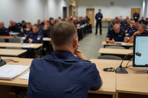 policjanci rozwiązują test siedząc przy ławkach. Obserwują ich funkcjonariusze pełniący rolę organizatorów zawodów