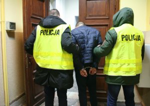 Dwaj policjanci w żółtych kamizelkach odblaskowych przy drzwiach wewnątrz budynku. Pośrodku młody mężczyzna z kajdankami z pochyloną do przodu głową