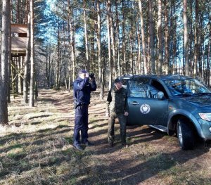 Droga leśna. Po środku policjant patrzy przez lornetkę. Obok z prawej strony o otwarte drzwi pojazdu należącego do Straży Leśnej opiera się Strażnik Leśny. W oddali w lewym górnym rogu widać ambonę myśliwską.