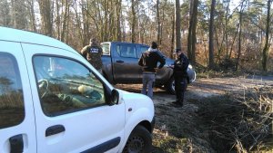 Z lewej strony zaparkowany biały samochód. Pośrodku stoi tyłem Strażnik Leśny oraz kontrolowany mężczyzna. Policjant spisuje jego dane w notatniku służbowym.