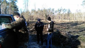 Z prawej strony policjant notuje w notatniku służbowym dane kierowcy, obok pośrodku kontrolowany mężczyzna. Z lewej strony obrócony bokiem Strażnik Leśny, który opiera się o samochód służbowy.