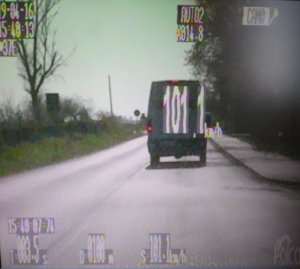 Pojazd typu bus koloru białego poruszający się drogą. W centralnej części fotografii widoczny wynik pomiaru prędkości - 101,1 km/h.