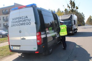 Policjant podczas czynności kontrolnych, z lewej strony zaparkowany samochód Inspekcji Transportu Drogowego, w oddali kontrolowany samochód ciężarowy i skrzyżowanie.