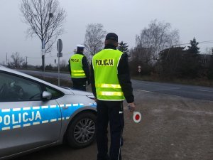 Dwaj policjanci ubrani w żółte kamizelki odblaskowe stoją przy radiowozie. Jeden z nich w ręku trzyma tarczę do zatrzymywania pojazdów. Obserwują nadjeżdżające pojazdy.