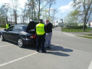 Policjanci zatrzymali do kontroli czarny samochód. Jeden z nich wspólnie z kierowca sprawdza zawartość bagażu przewożonego w bagażniku, natomiast drugi asekuruje.