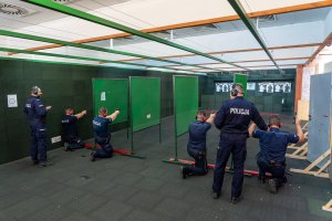 Policjanci na strzelnicy. Czterech dzielnicowych w pozycji klęczącej mierzy do tarcz strzeleckich. za ich plecami dwaj istruktorzy strzelań policyjnych.