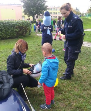 Przedszkolak wspólnie z mamą stoją z przodu radiowozu, oglądają kask ochronny używany podczas zabezpieczenia imprez masowych. Z lewej strony policjantka trzymająca w rękach kamizelkę odblaskową.