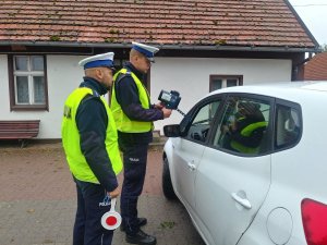 Policjanci ruchu drogowego ubrani w żółte kamizelki odblaskowe pokazują wynik pomiaru prędkości zarejestrowany przy użyciu laserowego miernika prędkości. Z lewej strony zaparkowany biały samochód.