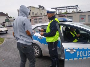 Policjant ruchu drogowego wręcza pieszemu opaskę odblaskową. W tle widać ulicę i przejeżdżające pojazdy. Z prawej strony w radiowozie znajduje się drugi policjant.