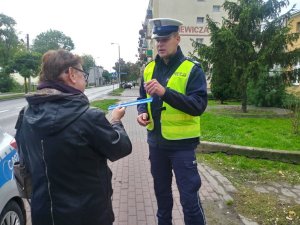 Policjant ruchu drogowego wręcza pieszej opaskę odblaskową zachęcając do jej noszenia.