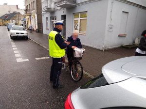 Policjant ruchu drogowego podczas rozmowy z rowerzystką. W tle widoczny chodnik i część ulicy z zaparkowanymi samochodami