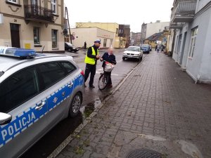 Policjant ruchu drogowego podczas legitymowania rowerzystki na jednej z ulic centrum Żnina. Z lewej strony widoczny zaparkowany radiowóz, natomiast po prawej stronie w oddali widoczna ulica z zaparkowanymi i przejeżdżającymi samochodami.