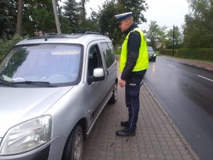 Policjant ruchu drogowego podczas kontroli drogowej stoi przy szarym pojeździe i prosi kierowce o okazanie dokumentów.