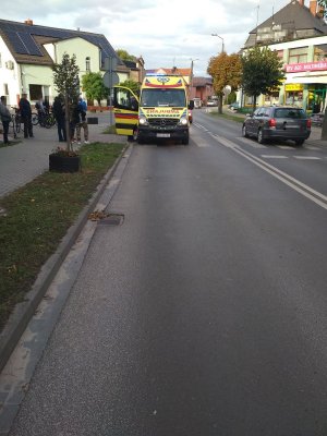 Widok ulicy, gdzie na przejściu dla pieszych stoi karetka pogotowia ratunkowego.  z Lewej strony widoczni są przechodnie, którzy przyglądają się całemu zajściu.