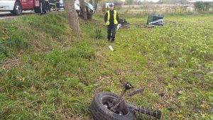 Części samochodowe rozrzucone po polu. W oddali policjant ruchu drogowego prowadzący oględziny.