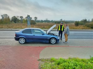 Policjant ruchu drogowego stoi z kierujących przed kontrolowanym pojazdem. W samochodzie jest podniesiona do góry pokrywa silnika.