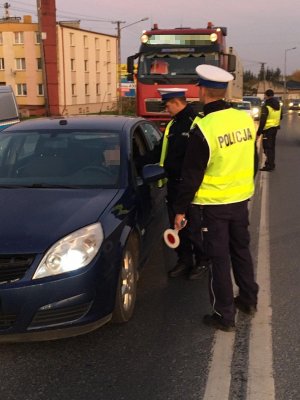 Policjanci ruchu drogowego stają na osi jezdni. Jeden z nich trzyma tarcze do zatrzymywania pojazdów. Drugi sprawdza stan trzeźwości kierowcy w granatowym samochodzie.