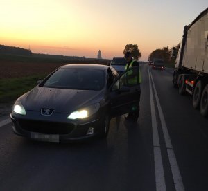 Policjant kontroluje kierującego w szarym samochodzie. W przeciwnym kierunku jedzie samochód ciężarowy.