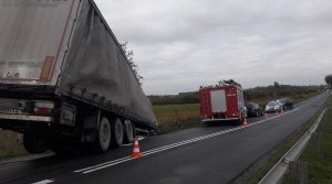 Fragment drogi wojewódzkiej. Z lewej strony widoczna naczepa pojazdu ciężarowego wystająca na lewy pas jezdni. Przed nią widoczne zaparkowane pojazdy m. in. straży pożarnej i policji.