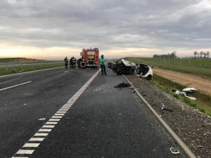 Fragment trasy S5, na który widoczny po prawej stronie rozbity biały samochód przy którym znajduje się technik kryminalistyki wykonujący dokumentację fotograficzną. Z lewej strony w oddali samochód Straży Pożarnej i strażacy zabezpieczający miejsce zdarzenia.