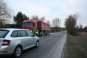 Fragment drogi dwukierunkowej. Na środku drogi stoją policjanci kontrolujący kierujących pod kątem trzeźwości. Z lewej strony srebrny samochód osobowy. Z prawej strony czerwony samochód ciężarowy z przyczepą.