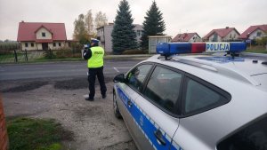 Policjant ruchu drogowego wykonuje pomiar prędkości przy użyciu laserowego miernika. Z prawej strony widoczny radiowóz.