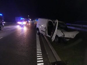 Fragment nowo budowanej drogi S-5. Na pierwszym planie uszkodzony samochód marki Fiat Doblo koloru białego. W tle panujący mrok. W oddali widać światła koloru niebieskiego nadawane przez pojazdy uprzywilejowane i funkcjonariuszy wykonujący czynności na miejscu wypadku.