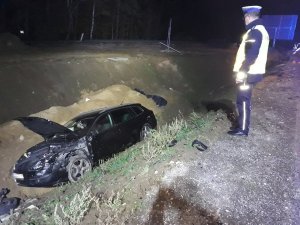 Funkcjonariusz ruchu drogowego, który ogląda uszkodzony samochód koloru czarnego leżący w przydrożnym rowie.