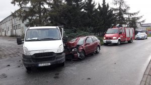 Z lewej strony widoczny biały samochód ciężarowy, w który z prawej strony uderzył samochód osobowy. W oddali z prawej strony widoczne pojazdy służb ratowniczych.