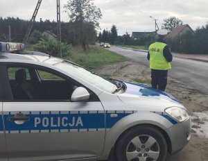 Policjant ruchu drogowego obserwuje ruch kołowy. Z lewej strony nadjeżdżają samochody. Obok stoi zaparkowany radiowóz.