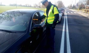 Policjant ruchu drogowego stoi na środku jezdni przy czarnym samochodzie osobowym. Sprawdza stan trzeźwości kierowcy przy użyciu alkomatu.