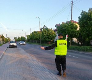 Policjant stoi na jezdni. Prawą rękę trzyma w górze z tarczą do zatrzymywania pojazdów. Lewą ręką wskazuje miejsce, w którym ma się zatrzymać pojazd.