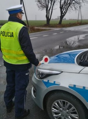 Policjant ruchu drogowego obserwuje ruch nadjeżdżających pojazdów. Z prawej strony widoczny radiowóz policyjny.