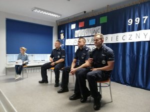 Policjanci z komisariatu Policji w Barcinie siedzą na scenie. Jeden z nich w dłoni trzyma mikrofon. Z lewej strony na krześle siedzi dziewczynka.