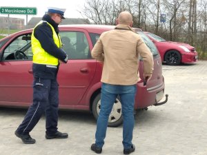 Funkcjonariusz ruchu drogowego podczas kontroli drogowej. Policjant wspólnie z kierującym stoją przed samochodem koloru czerwonego na utwardzonym placu.