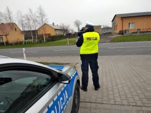 Policjant ruchu drogowego wykonuje pomiar prędkości przy użyciu laserowego miernika. Z lewej strony stoi zaparkowany radiowóz.