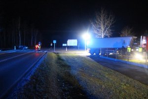 Widok miejsca zdarzenia. Skrzyżowanie, na którym na środku stoi strażak i kieruje ruchem. Z prawej strony widoczny budynek mieszkalny, a obok niego policjanci ruchu drogowego. Z Lewej strony na krawędzi lasu rozbity samochód osobowy. Panuje mrok.