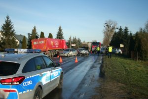 Miejsce zdarzenia drogowego. Z lewej strony na jezdni stoi radiowóz. W tle widoczne dwa uszkodzone samochody osobowe. Przy pojazdach stoją policjanci i strażacy, którzy wykonują czynności służbowe. Za nimi wahadłowo odbywa się ruch.