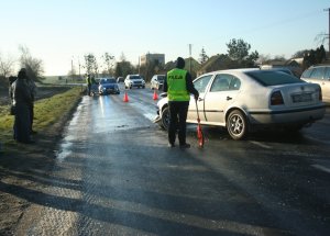 Technik kryminalistyki podczas wykonywania oględzin. W ręku trzyma kółko do wykonywania pomiarów odległości. Przed nim na pasie ruchu stoi samochoód koloru srebrnego. W oddali zaparkowany policyjny radiowóz.