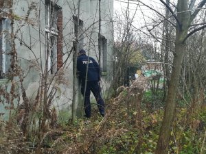 Zniszczony stary budynek. Szyby w oknach są powybijane. Przez jedno z nich zagląda policjant.