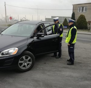 Z lewej strony fotografii znajdują się policjanci ruchu drogowego, którzy stoją przy czarnym samochodzie. kierowca znajduje się w swoim pojeździe.