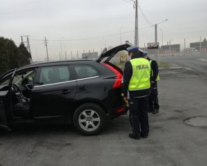 Policjanci podczas kontroli bagażu. Kierujący znajduje się za pojazdem, z bagażnika wyciąga wymagane przepisami wyposażenie.