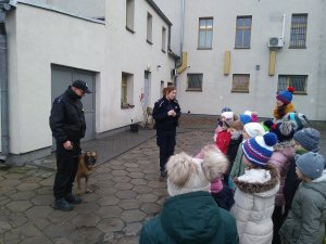 Policjantka wspólnie z dziećmi na dziedzińcu żnińskiej komendy. Opowiada dzieciom o przewodników psów służbowych. Z lewej strony przewodnik psa służbowego mł. asp. Dariusz Śliwiński ze swoim podopiecznym Nero - owczarkiem belgijskim  wyszkolonym do wyszukiwania zapachu narkotyków.