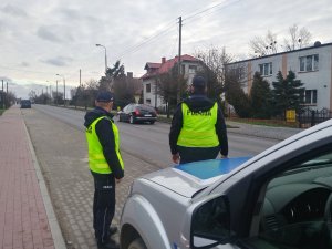 Policjanci obserwują ruch kołowy pojazdów. Za ich plecami stoi zaparkowany na poboczu drogi radiowóz.