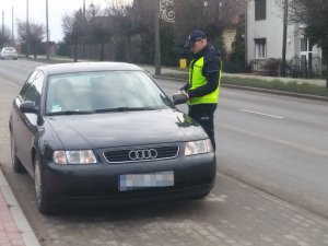 Policjant podczas kontroli samochodu osobowego. Prosi kierowcę, który siedzi w środku pojazdu o okazanie dokumentów.