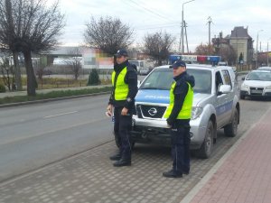 Policjanci obserwują ruch drogowy. Za ich plecami widoczny radiowóz. W oddali droga.