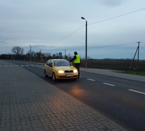 Policjant stoi na jezdni przy samochodzie osobowym koloru żółtego. Sprawdza kierowcę na obecność alkoholu w wydychanym powietrzu.