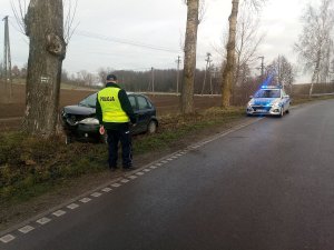 Policjant zabezpiecza miejsce zdarzenia drogowego. Funkcjonariusz stoi na lewym pasie jezdni. Przed nim w przydrożnym rowie znajduje się rozbity samochód.  w tle zaparkowany radiowóz.