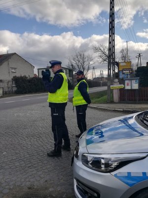 Policjanci podczas działań PRĘDKOŚĆ. Funkcjonariusze  stoją przy jezdni. Jeden z nich wykonuje pomiar prędkości. Z prawej strony policyjny radiowóz.