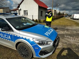 Policjant ruchu drogowego wykonuje pomiar prędkości. Z lewej strony radiowóz zaparkowany na poboczu drogi.